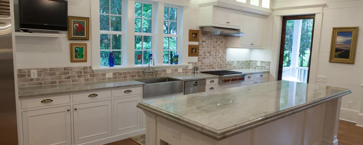 White custom kitchen with a large marble-topped island, farmhouse sink, brick backsplash and tall windows